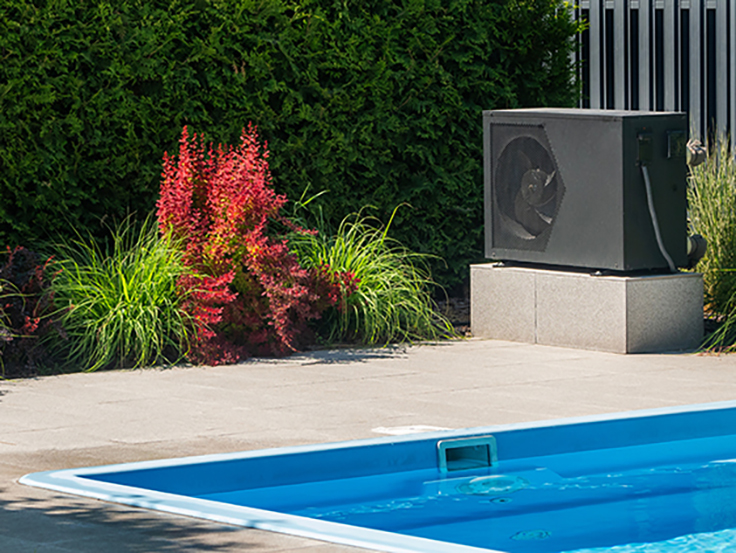 An outdoor unit of a heat pump is installed at the edge of a swimming pool. The unit is black and is placed on a sturdy concrete platform.
