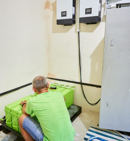 Technician working on green electrical battery storage unit placed on black base on floor. On the wall two modern inverter units connected to battery by black cables. Man installing solar panel system
