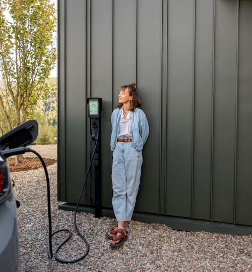Woman waits for electric car to be charged standing near a charging station leaning to a house wall. Concept of modern lifestyle and smart technologies